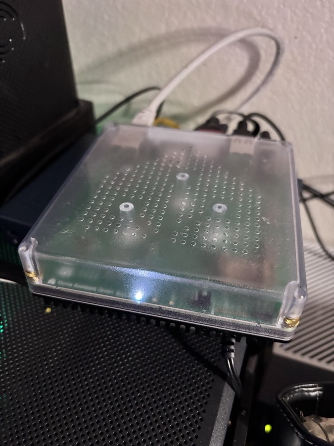 The Home Assistant Green, a small box with a clear lid and a blue status light, on a shelf in the office