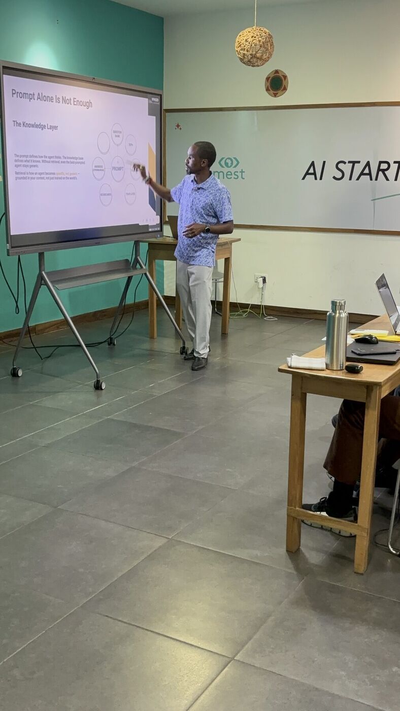 Waziri Garuba at the front of the MEST classroom in Accra, pointing at a slide, with the MEST logo and the words AI Startup Program on the wall behind him.