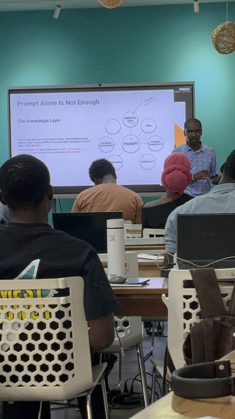 Waziri at the screen pointing to a slide titled The Knowledge Layer: a prompt at the center, ringed by a question bank, SOPs, founder transcripts, templates, scorecards, and strong and weak answers.